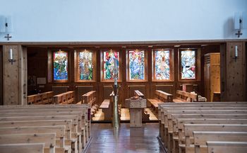 Das Bild zeigt den Innenraum einer Kirche. Es sind Sitzbänke aus Arvenholz zu sehen. Im Hintergrund sind bunte Kirchenfenster zu sehen.
