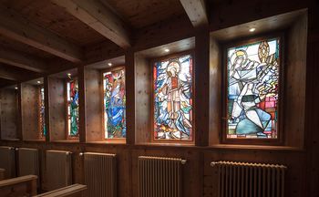 Das Bild zeigt mehrere bunte Kirchenfenster. Unter jedem Fenster ist ein Heizkörper an der Wand angebracht.