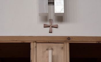 Das Bild zeigt eine weisse Stabkerze auf einem gusseisernen Kerzenständer. Der Kerzenständer ist an einer Säule aus Arvenholz befestigt.